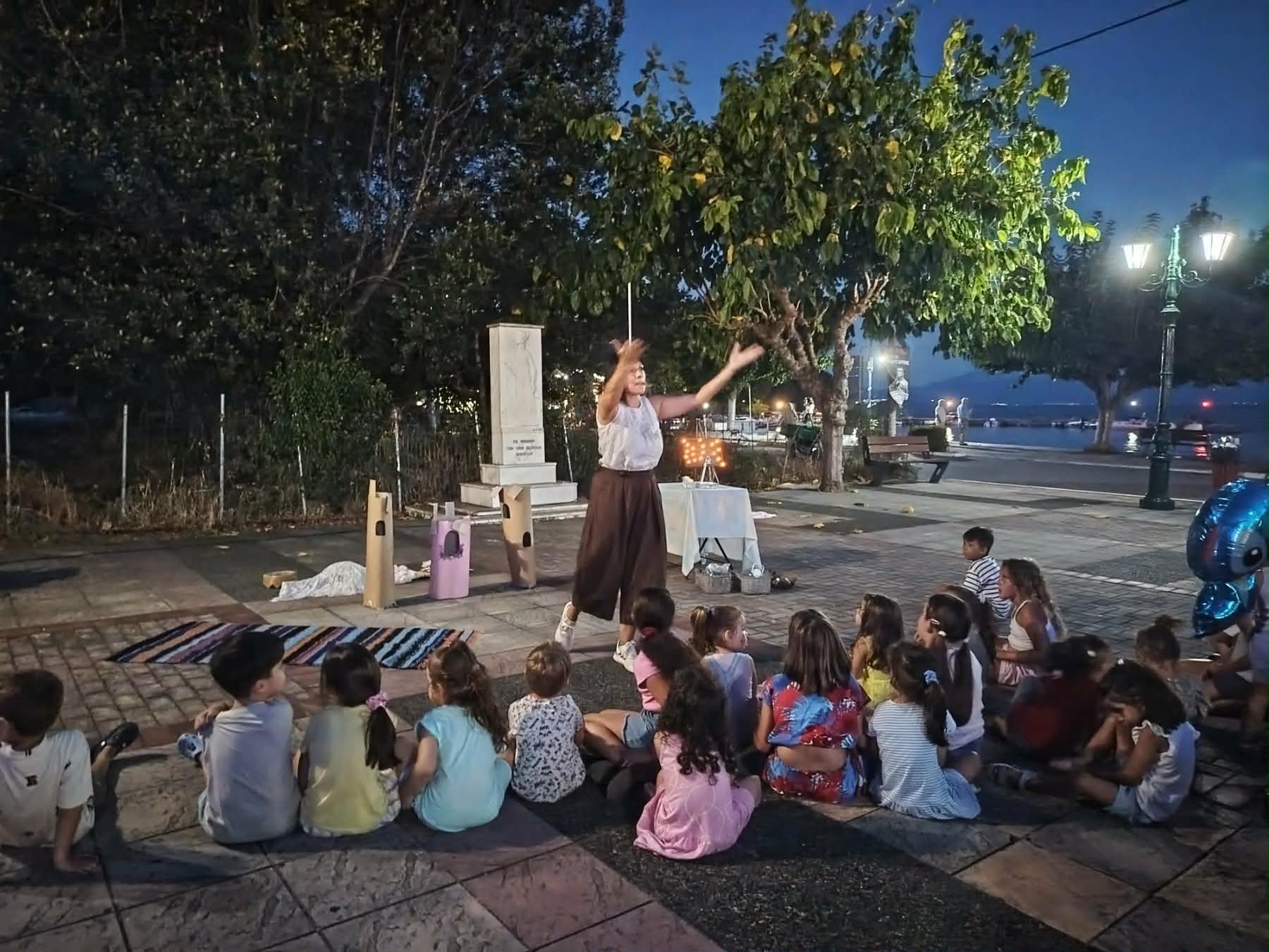 Η Παναγιώτα Στραγανιώτη αφηγείται σε παιδιά σε υπαίθριο χώρο δίπλα στη θάλασσα, το βράδυ