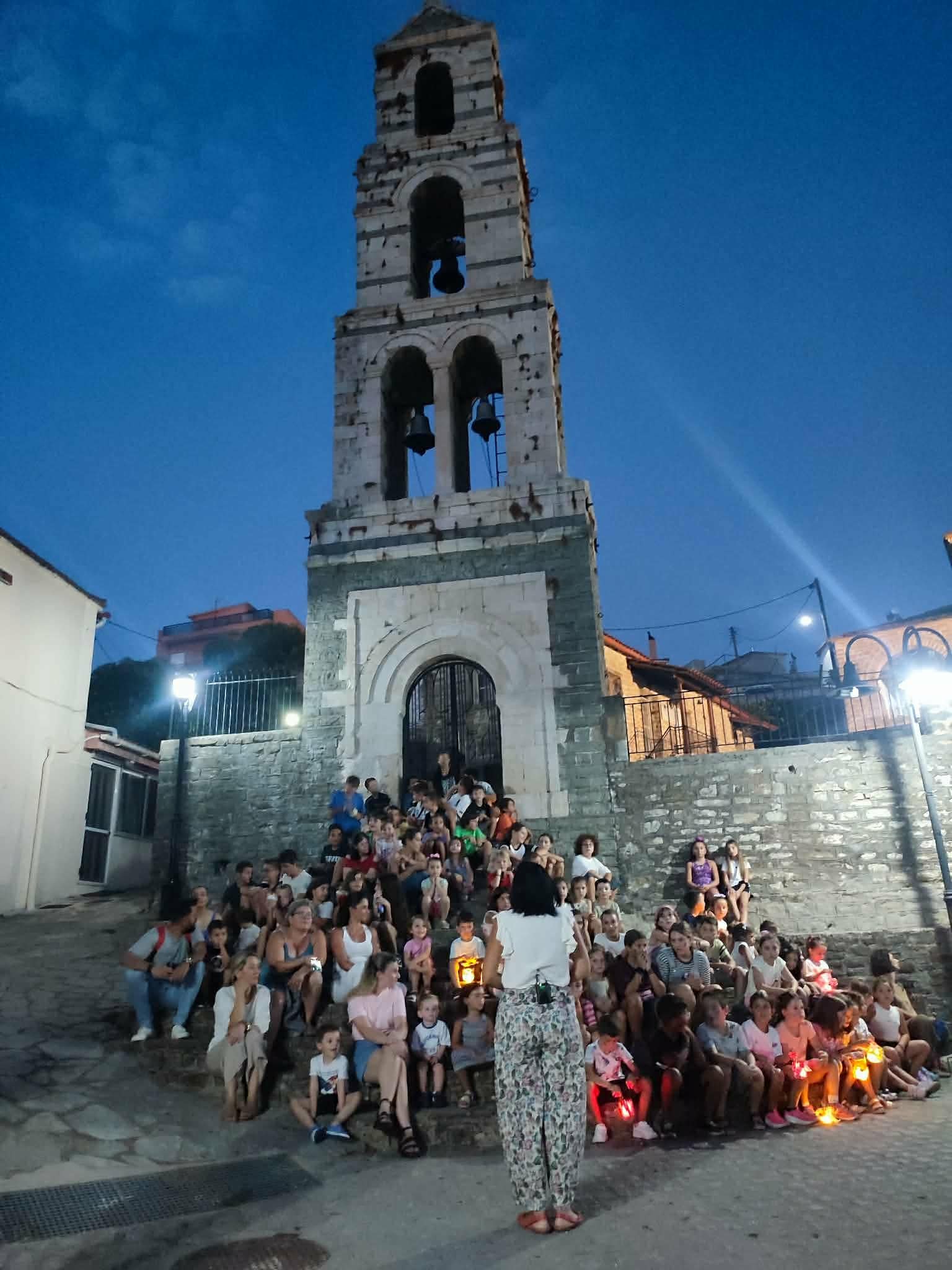 Παιδιά παρακολουθούν βραδινή αφήγηση της Παναγιώτας Στραγανιώτη έξω από παλιό καμπαναριό, κρατώντας φαναράκια